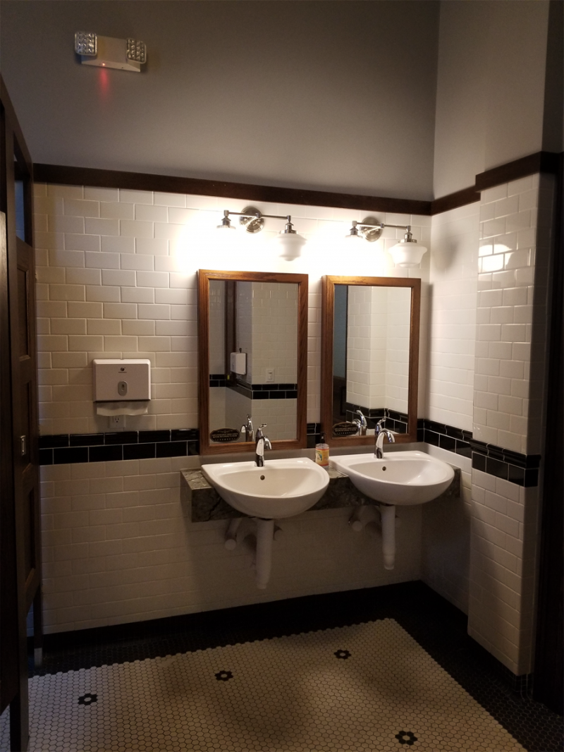 Two white porcelain sinks are side by side, each with a rectangular mirror above it and two lights connected by a thin bar. The wall is tiled with white brick-shaped tiles. The floor is blue and white mosaic tiled. Photo by Andy Long. .  
