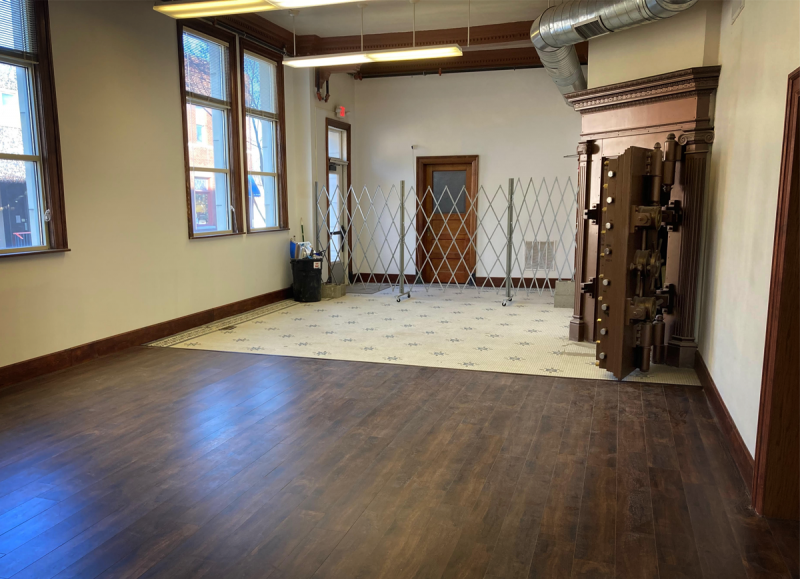 A vacant room. In the foreground is wood flooring. The back of the room has blue and white mosaic tiling. A metal divider stretches across the room. To the right is the door to a vault. There are two windows on the left side wall. Photo by Andy Long.