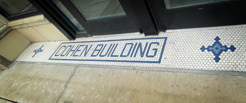 A rectangular section of tiling on the ground in front of a door. It's mostly white with blue accents. Blue tiles spell out COHEN BUILDING, with a thin blue border. Photo by Andy Long.