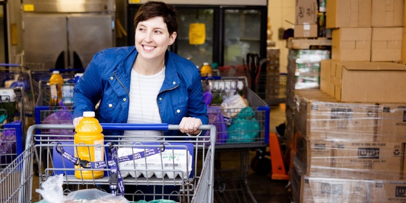 A white woman with short brown hair is pushing a shopping cart through a warehouse. She is wearing a blue jacket with a white shirt underneath.