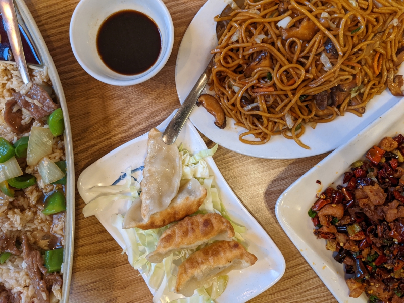 On a brown table, there are several Chinese dishes. On the far left, there is a long dish of beef and veggies. In the center up top is a small bowl of soy sauce, and below there is a plate of fried dumplings. On the right up top is a noodle dish and the right bottom is a dry chili chicken dish. Photo by Tayler Neumann.