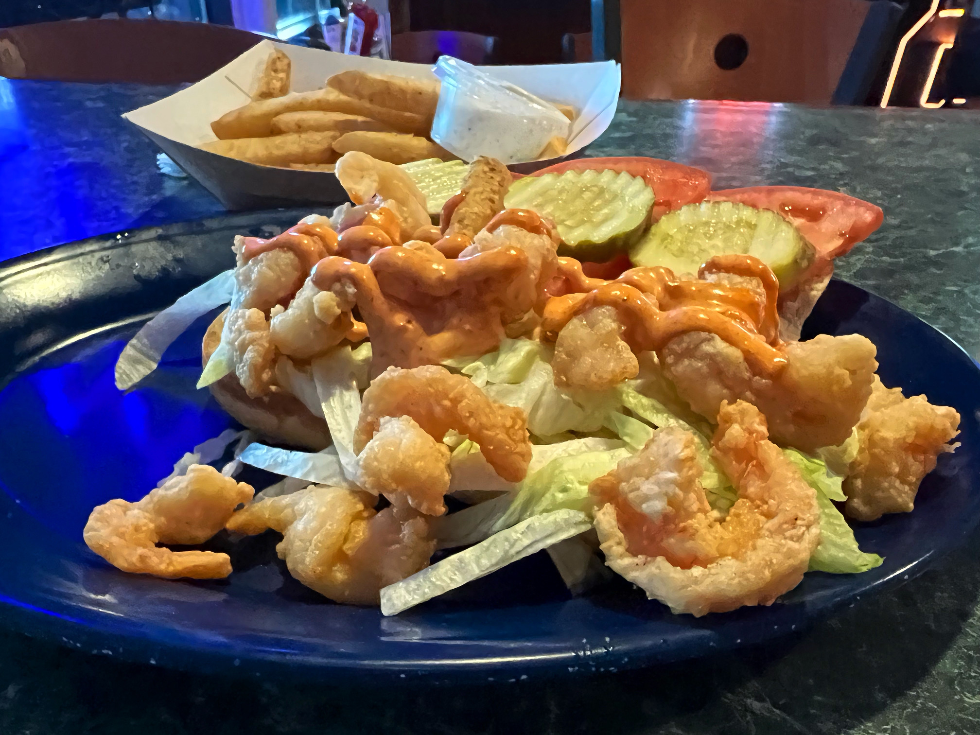 On a dark blue plate, there is a po boy with lots of shrimp and toppings. Some fried shrimp and shredded lettuce are on the plate, off of the sandwich. Photo by Alyssa Buckley.