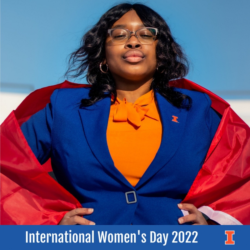 A Black woman with glasses and medium length dark wavy hair stands with her hands on her hips. She is wearing a blue blazer and orange blouse, with a Block I pin on her lapel. Photo from Women and Gender in  Global Perspectives Facebook page.