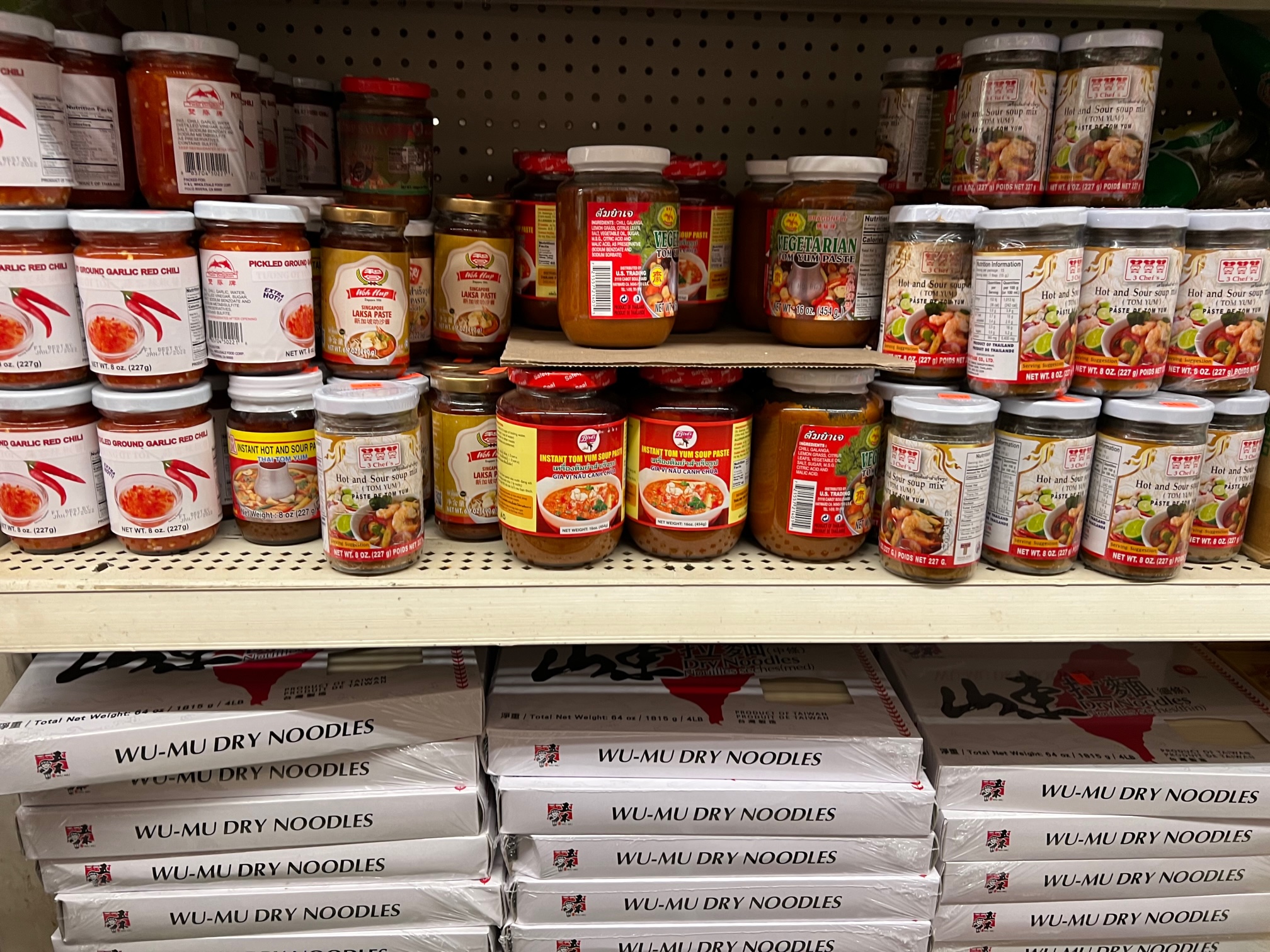 Two shelves at Far East Grocery show jars of sauces with peppers, and on the bottom, there are boxes of wu-mu dry noodles. Photo by Alyssa Buckley.