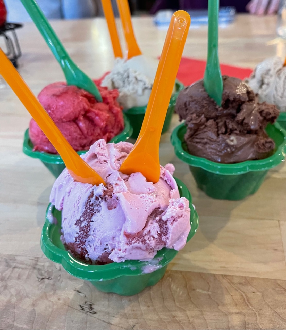 On a light wood table, there are six green cups of gelato of various flavors with plastic green spoons sticking out of them. Photo by Stephanie Wheatley.