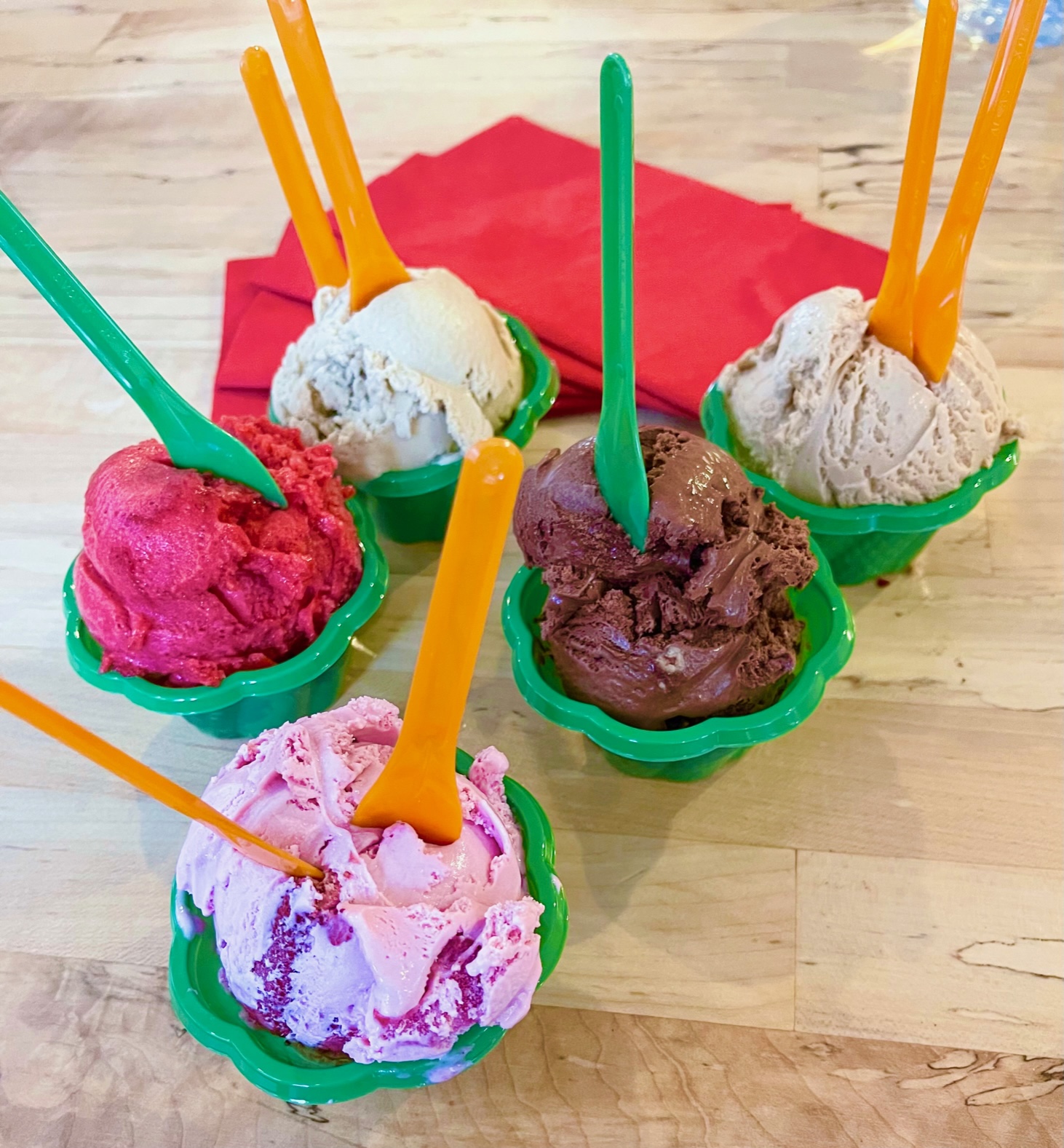 On a light wood table, there are five cups of gelato in green cups with various flavors and differently colored plastic spoons in each cup. Photo by Stephanie Wheatley.