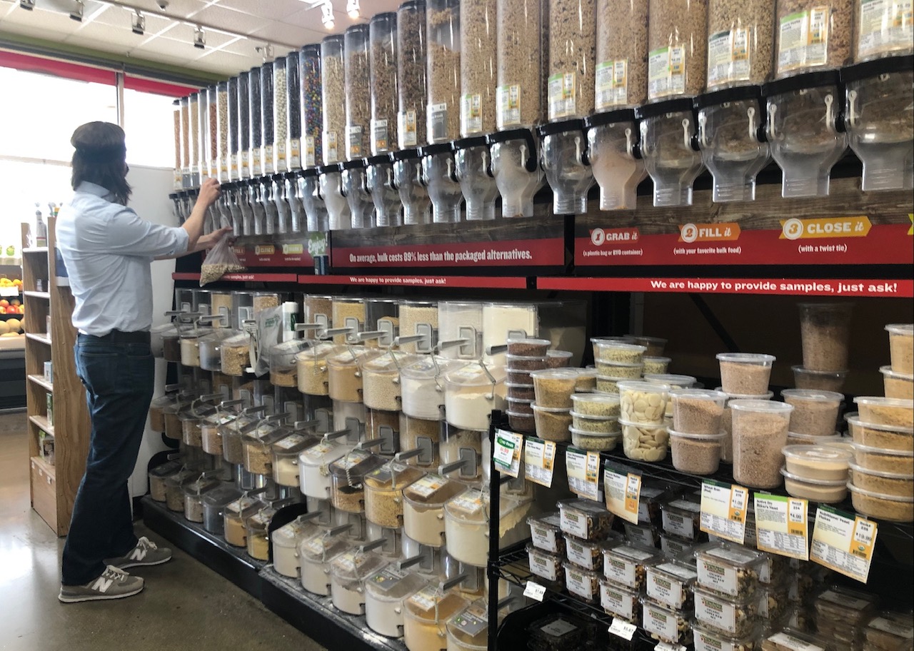 In the bulk aisle of Common Ground, there are rows of bulk foods. There is a white man facing away, serving some snacks into a plastic bag. Photo by Alyssa Buckley.