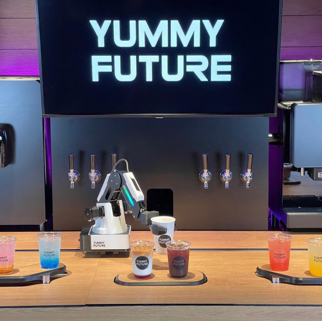 On a wooden counter, there is a small white robot with a very long arm gripping a white paper coffee cup. On the three trays on the table are a variety of other colorful drinks in clear to go cups. Photo from Yummy Future's Instagram. 