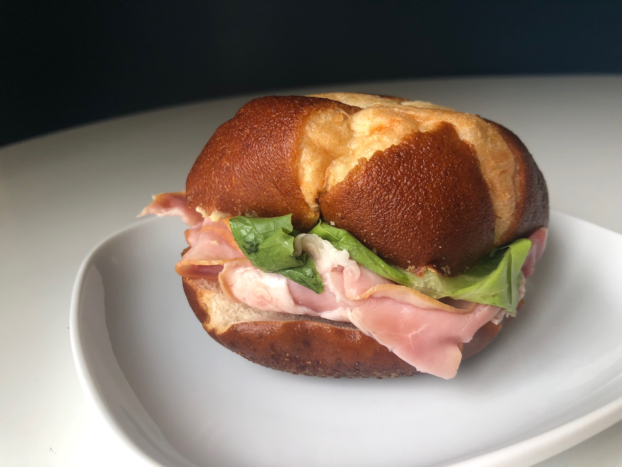 On a small white plate, there is a ham sandwich on a pretzel roll. Photo by Alyssa Buckley.