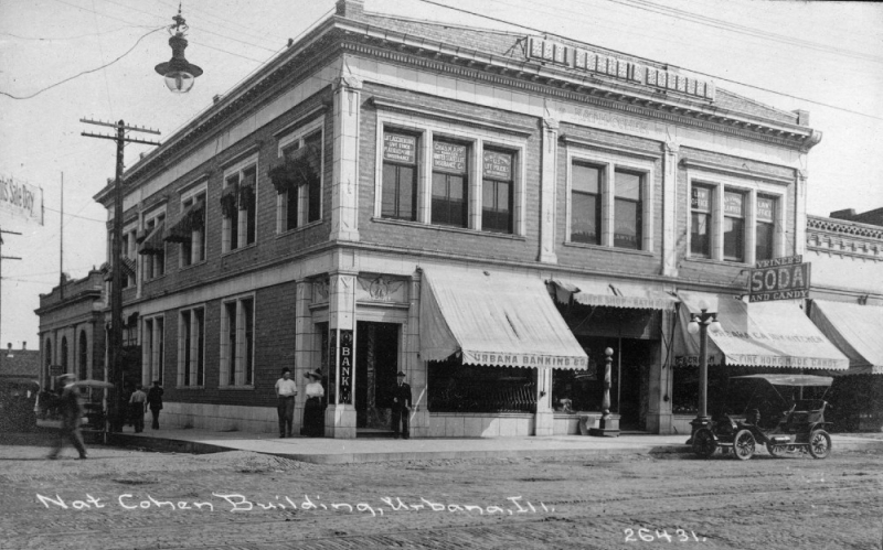 A black and white photo of a two story building on a street corner. The facade of a two story building. There are storefronts with large windows along the first floor, and rectangular windows framed by brick along the second floor. There is a large awning stretched over one of the first floor windows. Photo from The Cohen Building website. 
