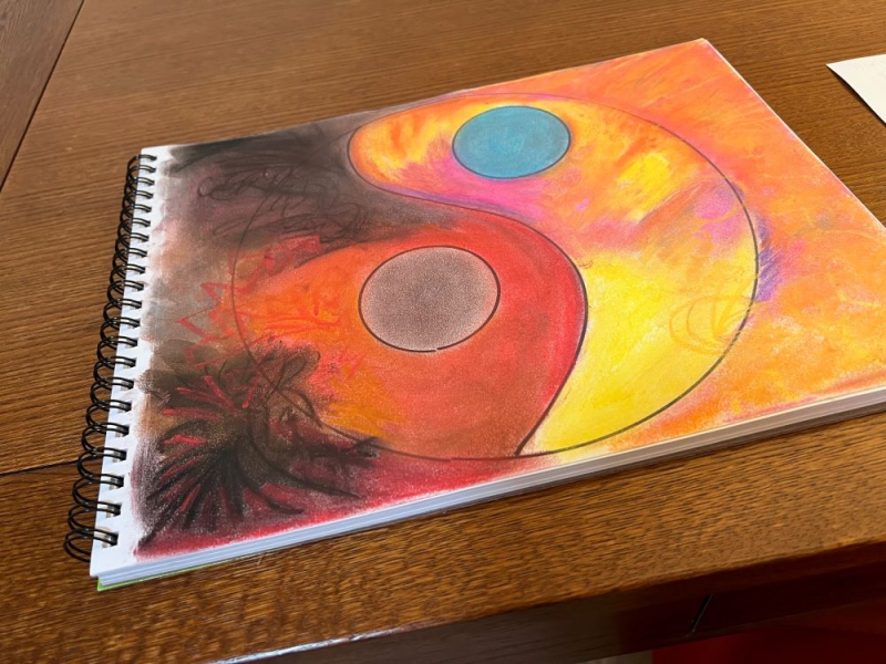 A spiral bound sketchbook sits on a wooden table. The page showing has a yin and yang symbol, and has been covered in color with pastels. In the corners are black shading. Photo by Julie McClure.