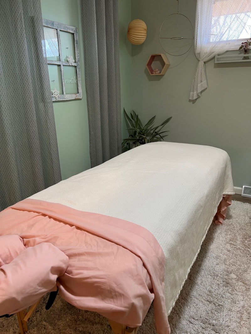 A massage table with a white blanket and pink sheets is the the middle of a small room with light green walls and curtains. Photo by Julie McClure.