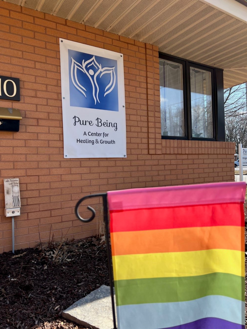 A corner of the exterior of a brick building. There is a white sign with a blue and white logo that says Pure Being, A Center for Healing and Growth on the side of the building. In the foreground there is a rainbow flag on a black wire stake in the ground. Photo by Julie McClure.