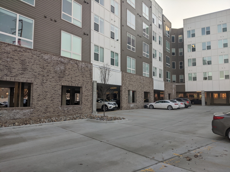 Two apartment buildings in a complex frame a parking lot. There is brick along the bottom, then alternating white and gray siding, with columns of windows. A few cars are parked in the lot. Photo by Tom Ackerman.