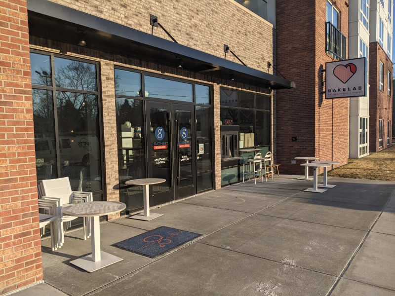 The entrance to a cafe. There are glass doors in the center, framed by tall windows. A few small tables and chairs are scattered on the cement patio, and a sign that says BakeLab is in front of the building. Photo by Tom Ackerman.