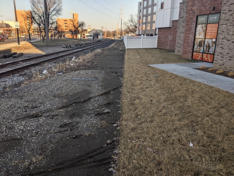 A railroad track runs behind an apartment building. It's surrounded by dirt, and there are a few bare trees in the background. Photo by Tom Ackerman.