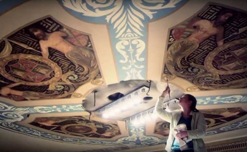 A woman in a gray cardigan is standing just below an ornately painted ceiling. She's holding a bucket in one hand and is reaching up towards the ceiling with a paintbrush. Screenshot from Shatterglass Studios.