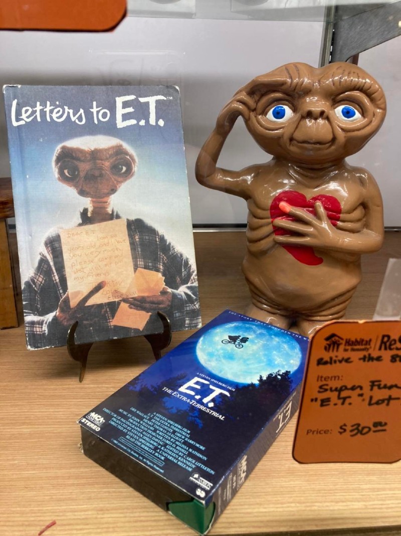 A figure of E.T. (brown alien with a large head and eyes and a red heart painted on its chest) sits on a wooden table next to a book with the image of E.T. on it, and a VHS cassette tape of the movie. It has a blue cover and the image of a moon with a bicycle flying across it. Photo from Champaign County ReStore Facebook page. 