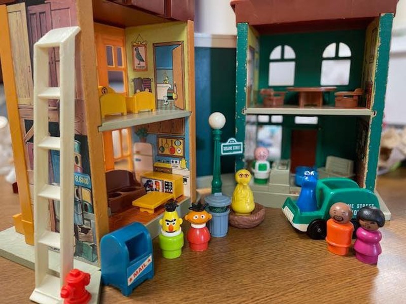 A vintage playset that is opened along a hinge to show two sides: the inside of an apartment with miniature furniture, and the inside of a grocery store. There are several Sesame Street character figures set in front of the playset. Photo from Champaign County ReStore Facebook page.