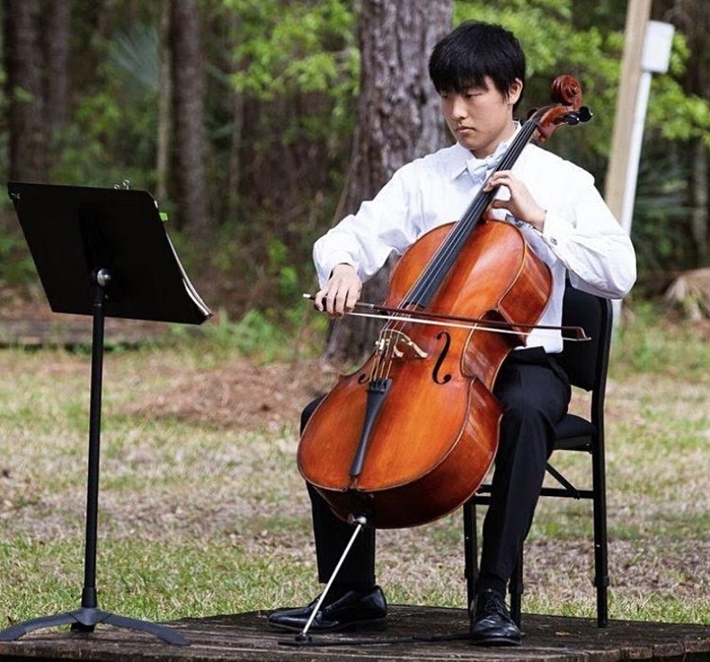 A Japanese man in black trousers and a white button down shirt is sitting in a chair playing a cello. There is a music stand in front of him, and he is in the middle of a grassy field with trees in the background. Photo provided by Japan House.