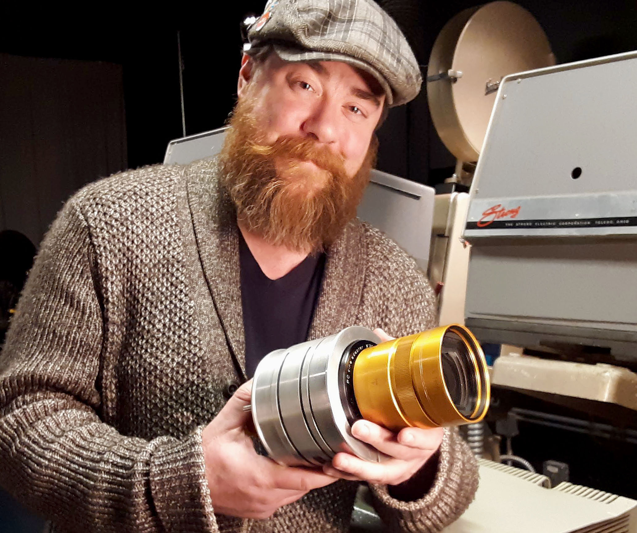 A man with a long red beard wearing a black hat and gray cardigan is holding a cylindrical lens in his hands. He is standing in front of film projection machine. Photo by Paul Young.