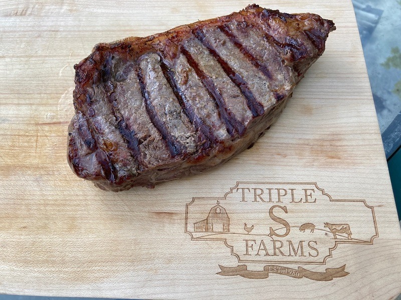 On a light colored wooden cutting board, there is a large piece of meat, grilled to perfection. In the bottom right corner of the cutting board, there is a black inscription of 