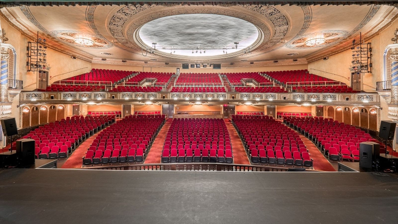 A view of a theater from the stage. There are rows of red seats on the main floor and in a balcony. The seats sit under an ornate ceiling. Photo by 3D Tour Gallery Photography.