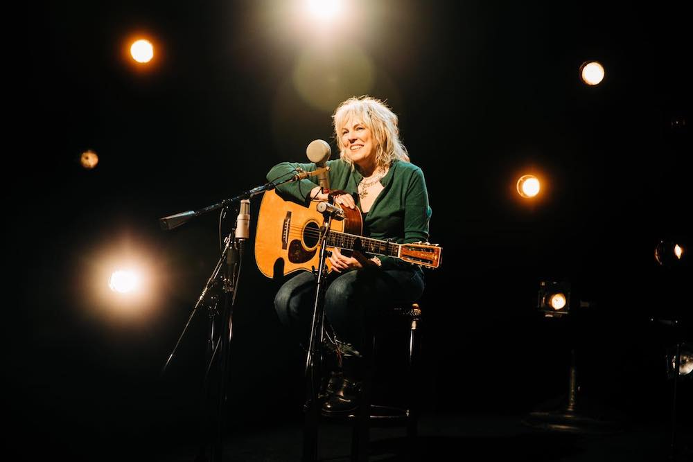 Portrait of Lucinda Williams sitting on a chair on stage. She is smiling bright, and holding a guitar in her lap. 