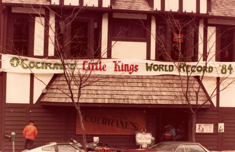 The exterior of a bar with an awning with wooden shingles. A banner is stretched across which says Cochrane's Little Kings World Record '84. Photo from Champaign Urbana History Facebook group.