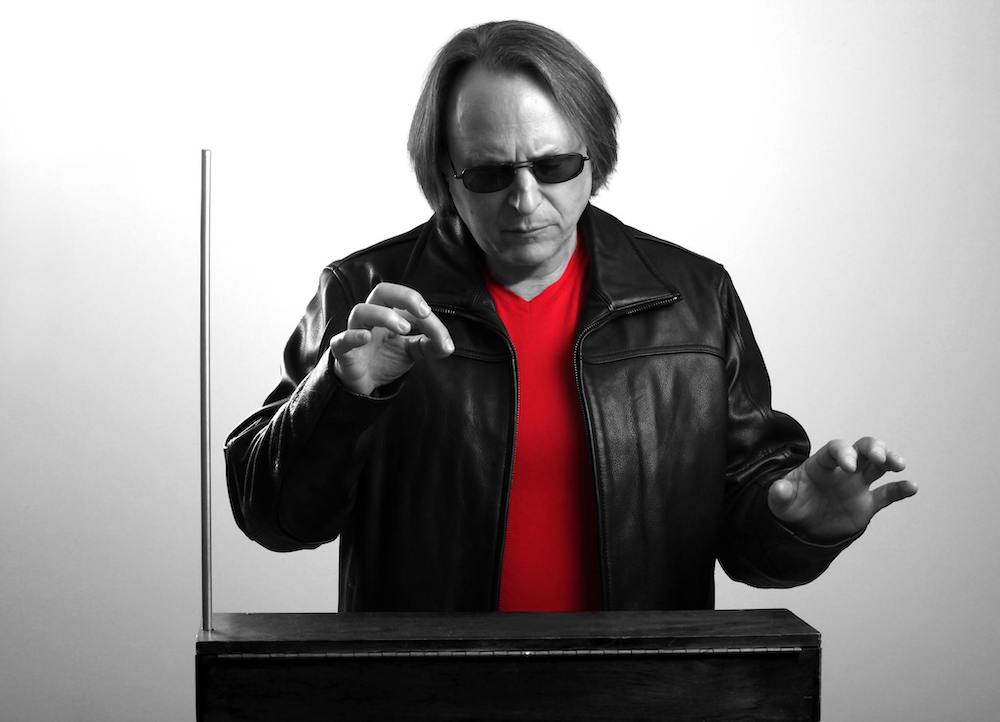 A black and white portrait of Rob Schwimmer playing the theremin, except for his shirt, which is a bright red. 