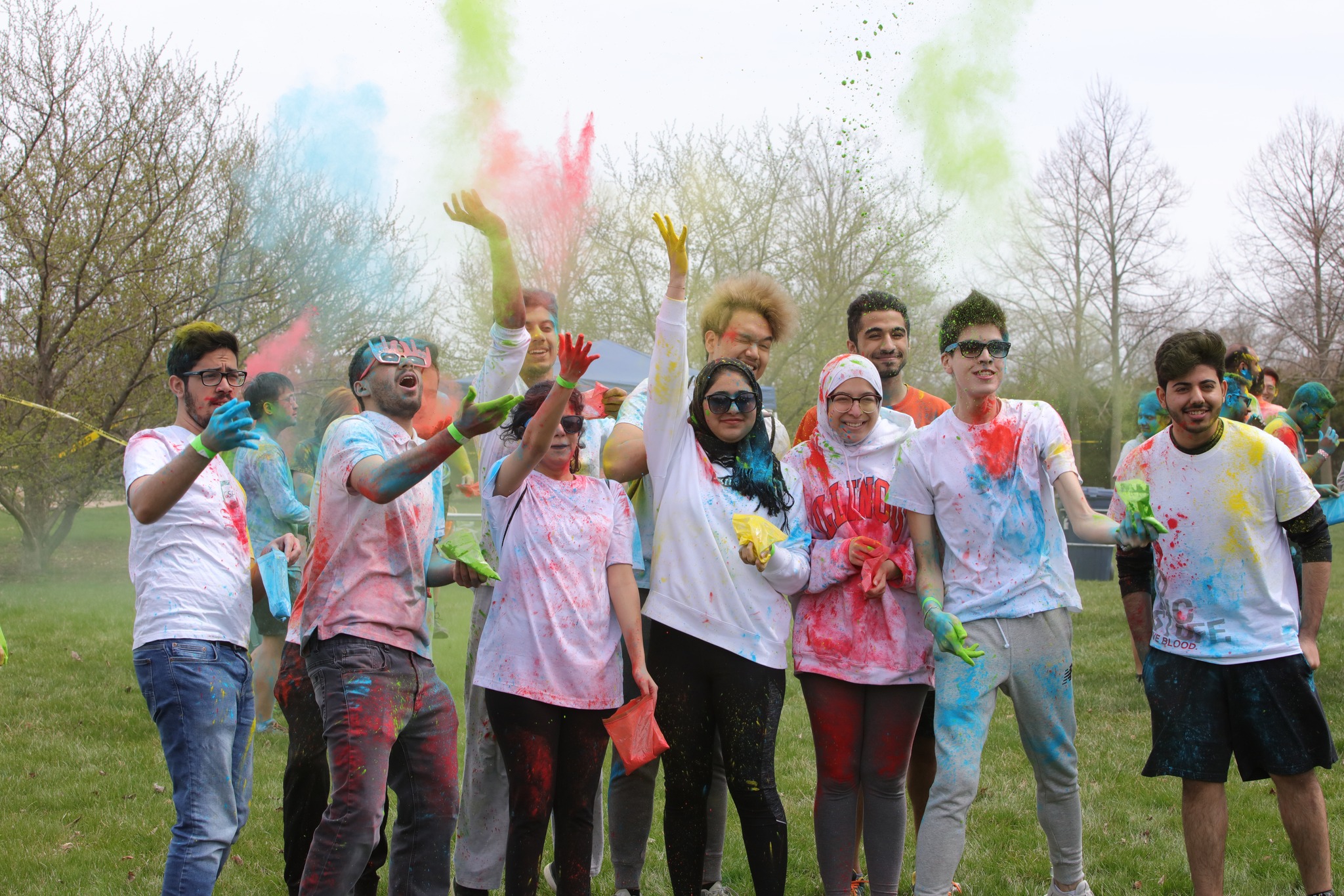 A group of young people in white t-shirts celebrate Holi. Their shirts are stained with colorful paint, and they are all throwing colorful paint/powder into the air. Photo from the Asha for Education â€” UIUC Facebook page.