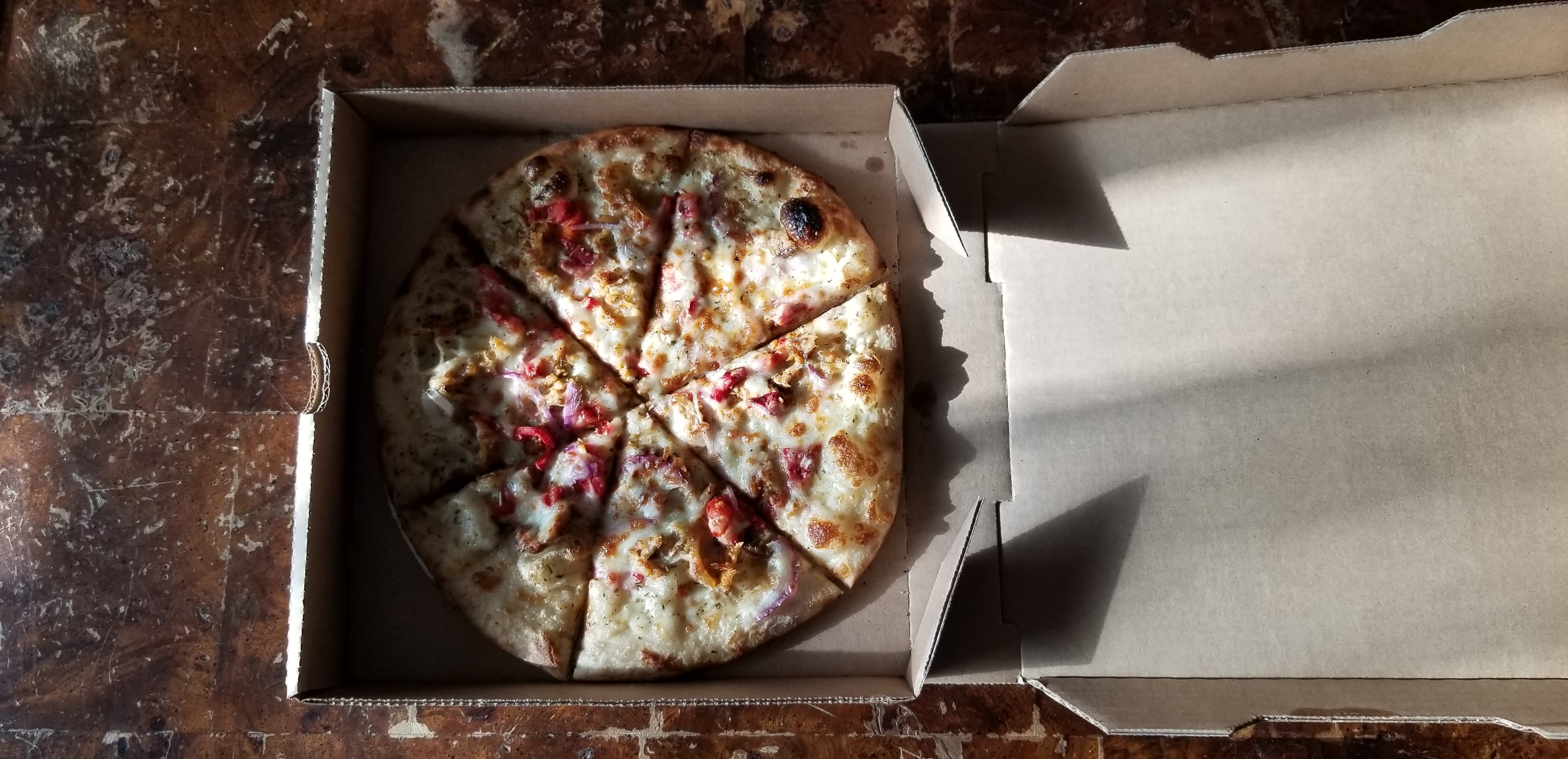 On a sun-drenched table, there is an open cardboard pizza box with a full BYO pizza sliced. Photo by Nina Hopkins.