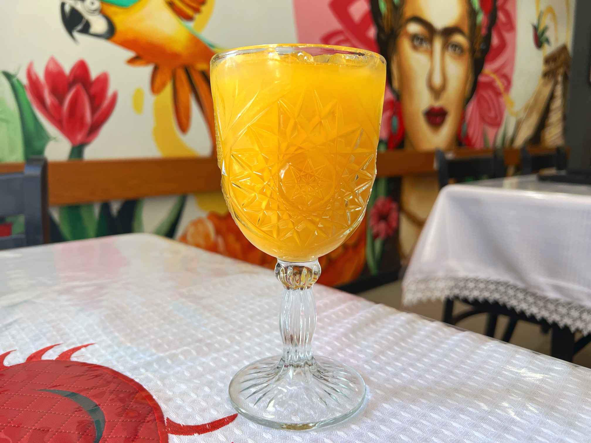 On a white tableclothed table, there is a glass goblet with an orange mango margarita. In the background, there are two colorful murals on La Bahia Grill's dining room walls. Photo by Alyssa Buckley.