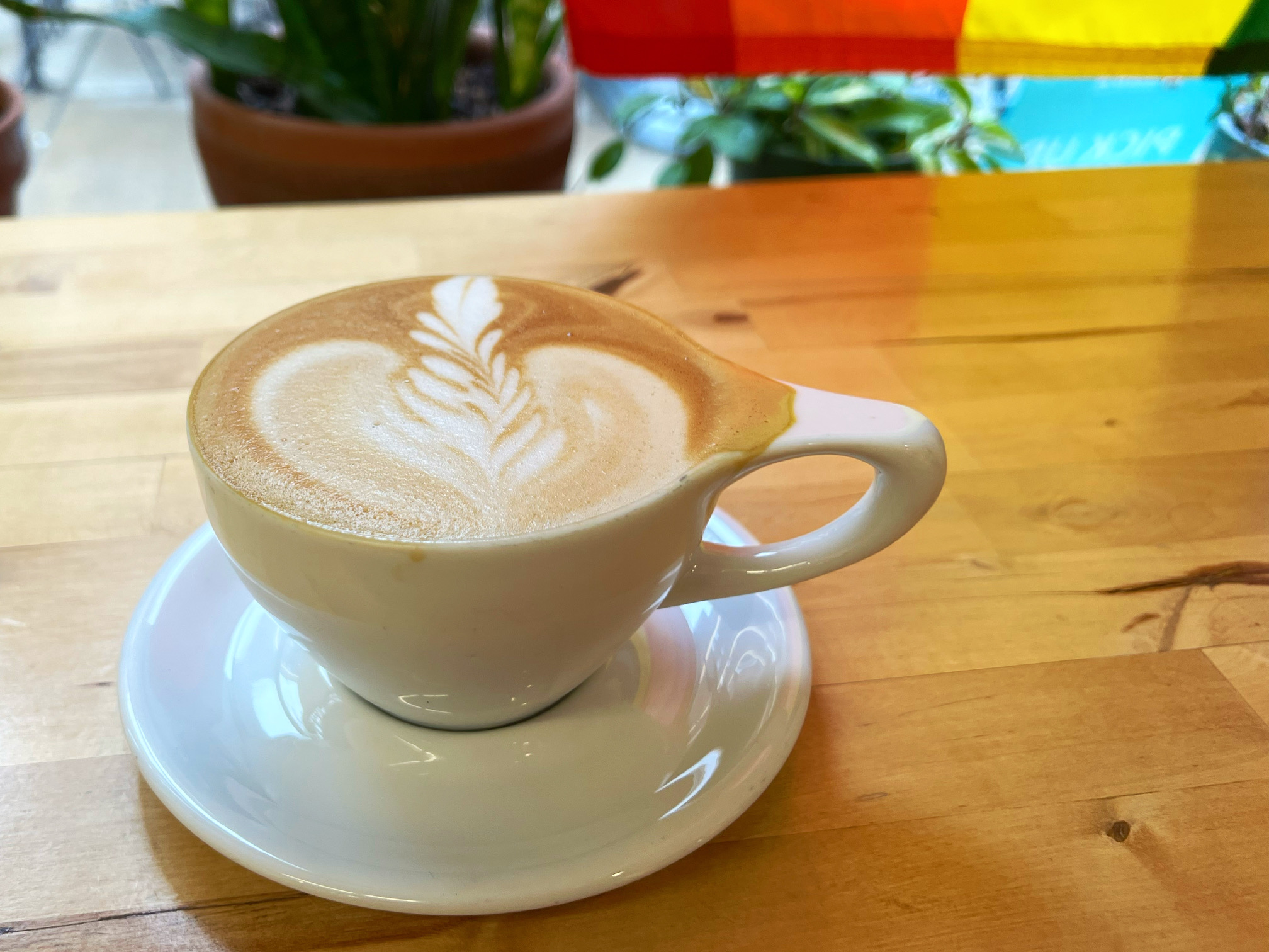 On a light wooden counter, there is a white coffee cup with a white saucer beneath it. The latte has an artfully made latte art on top. Photo by Alyssa Buckley.