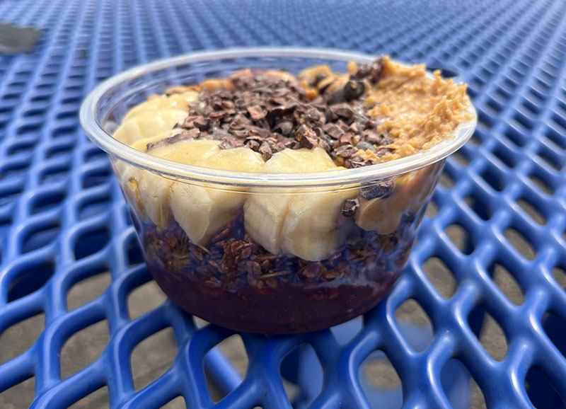 A clear plastic bowl sits on a blue mesh picnic table. A layer of acai fills the bottom of the bowl. Banana slices, peanut butter, and cacao nibs are visible on top. Photo by Julia Freeman.