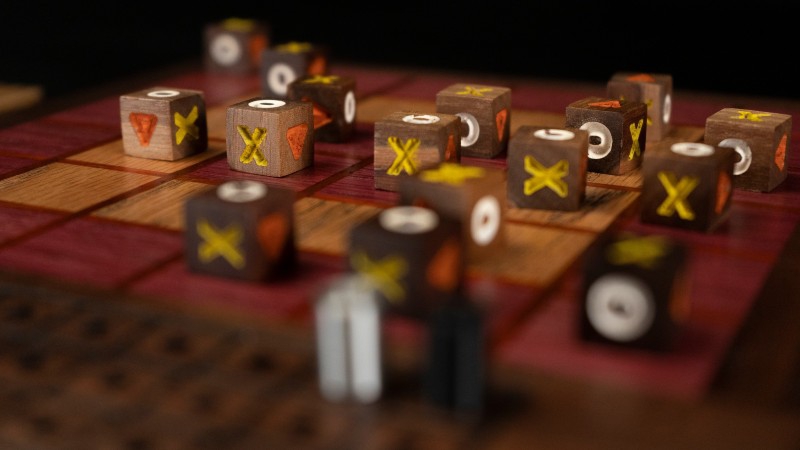A close up of several wooden cube game pieces on a wooden board with squares. The cubes have different symbols on each side: X, O, triangle. Photo from CUDO Plays Facebook page.