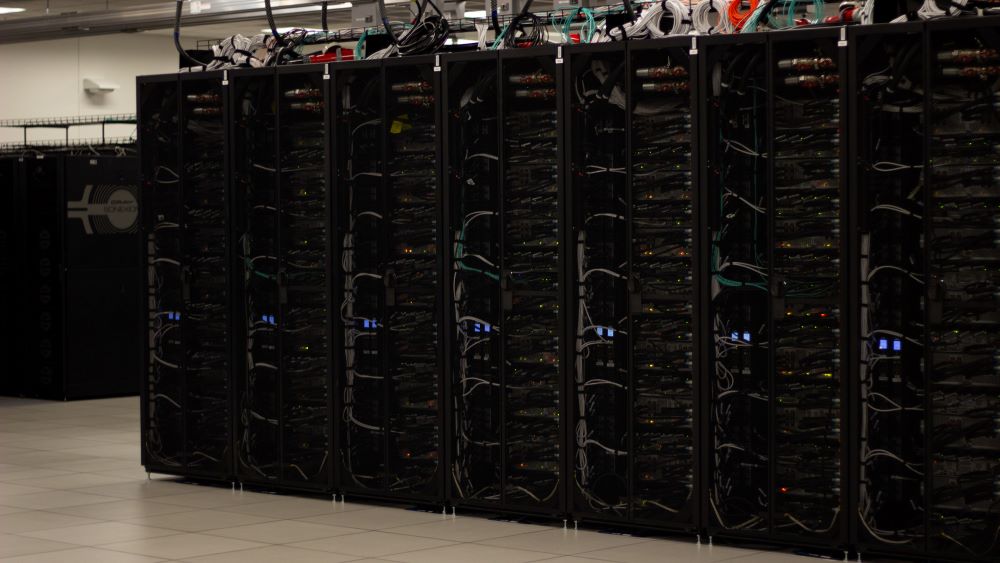 A row of tall black supercomputer components. They have multicolored wires on top of them.