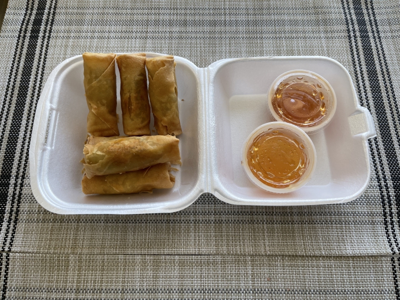 There are egg rolls in a small square styrofoam container with a small cup of red sauce. Photo by Shrivatsa Ravikumar.