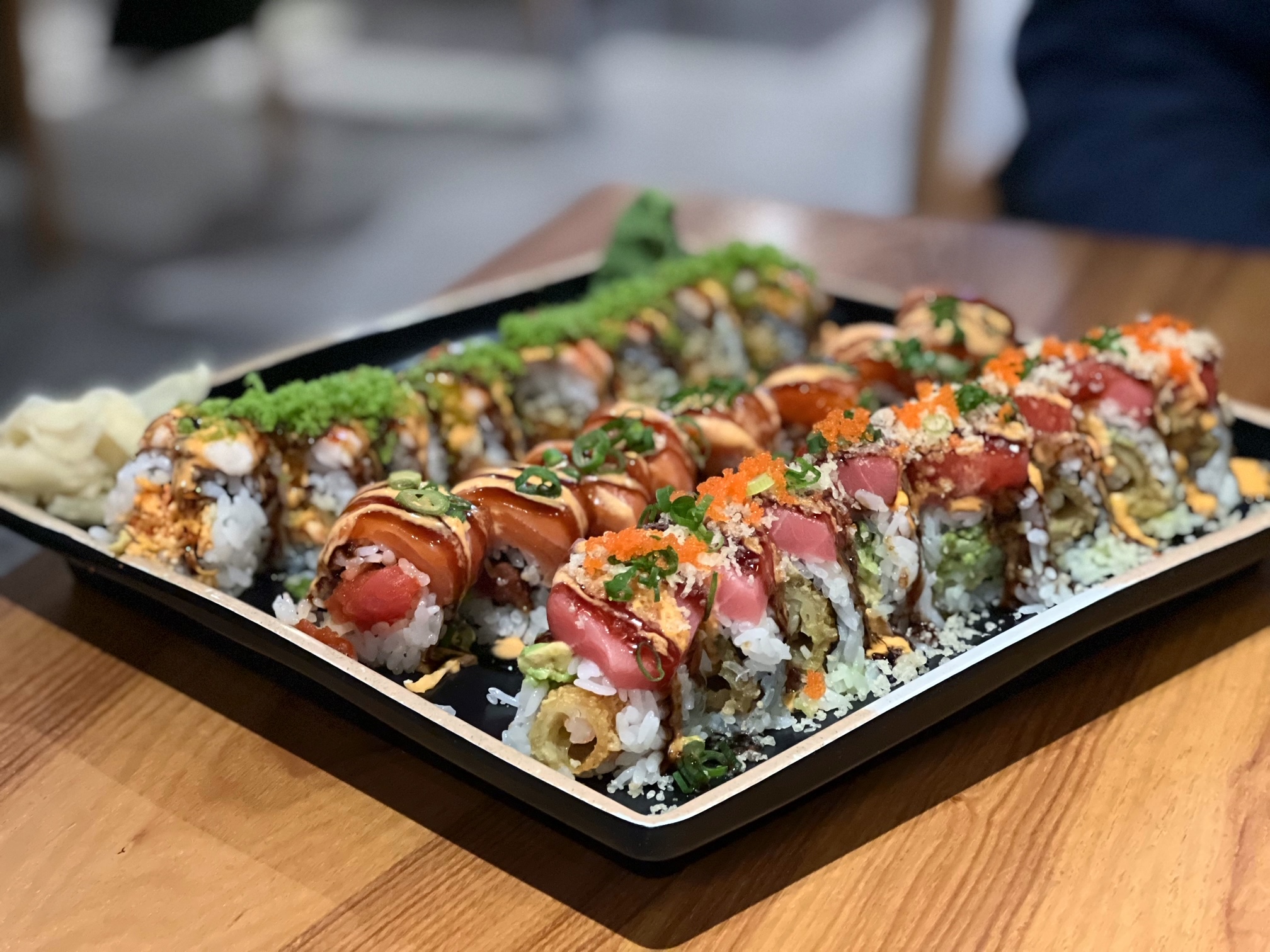 On a wooden table, there are three sushi rolls with various fish, sauces, colorful crunchies, and flakes. Photo by Alyssa Buckley.