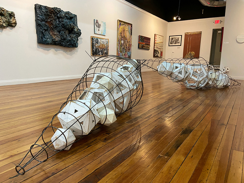 Geometric wire and wood sculpture by Nathan Westerman in the foreground with paintings lining the walls behind at Gallerie 112.