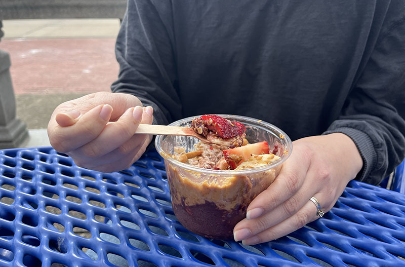 A woman in a black, long-sleeved t-shirt sits at a blue mesh picnic table. She holds a clear plastic bowl with peanut butter, aÃ§aÃ­, granola, and fruit. She lifts a strawberry slice with a wooden spoon. Photo by Julia Freeman.