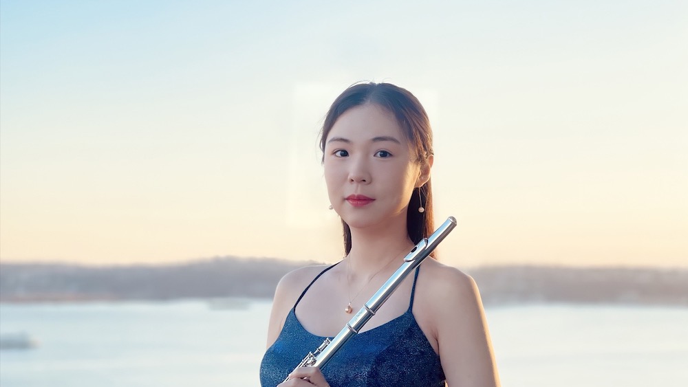 Portrait of Peiyao Cheng holding her flute, a blue sky and body of water behind her.