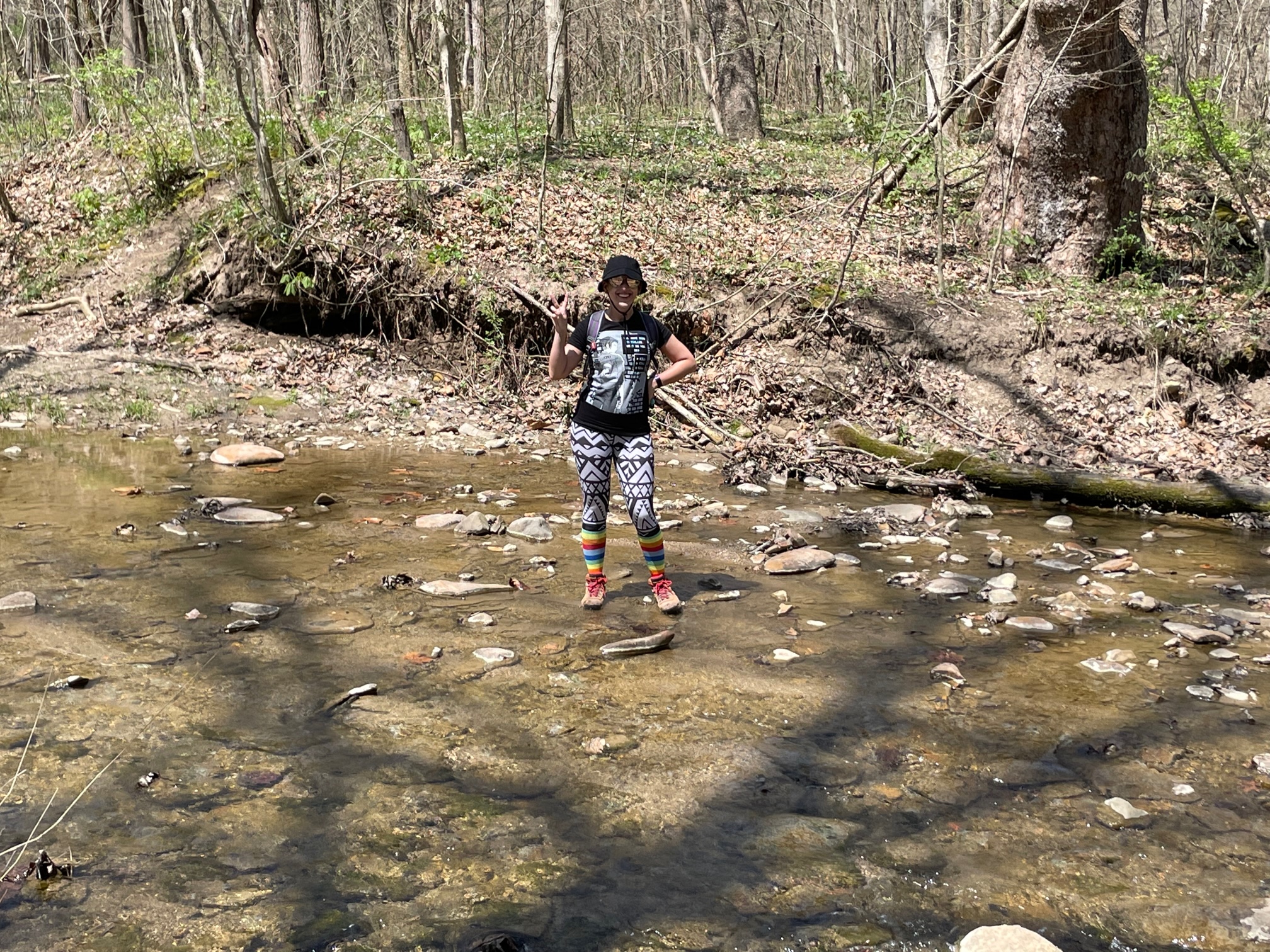 Photo of author Mara Thacker standing in the middle of a shallow stream with rocks. There are trees in the background, main colors in the are browns and greens.