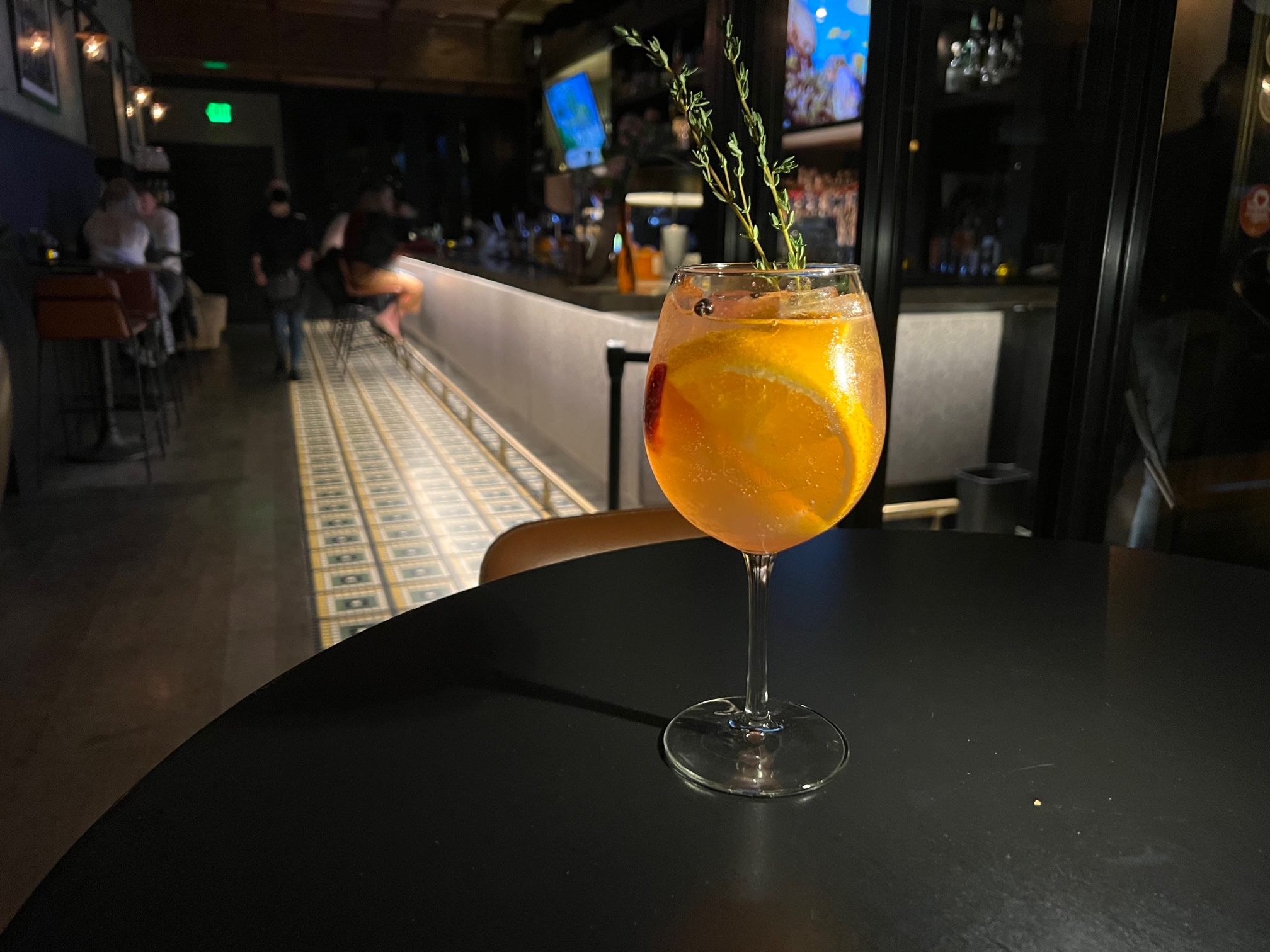 In a dimly lit bar, there is an orange cocktail in a wine glass on a circular black table. The glass has orange slices, strawberries, and springs of thyme in it. Photo by Alyssa Buckley.