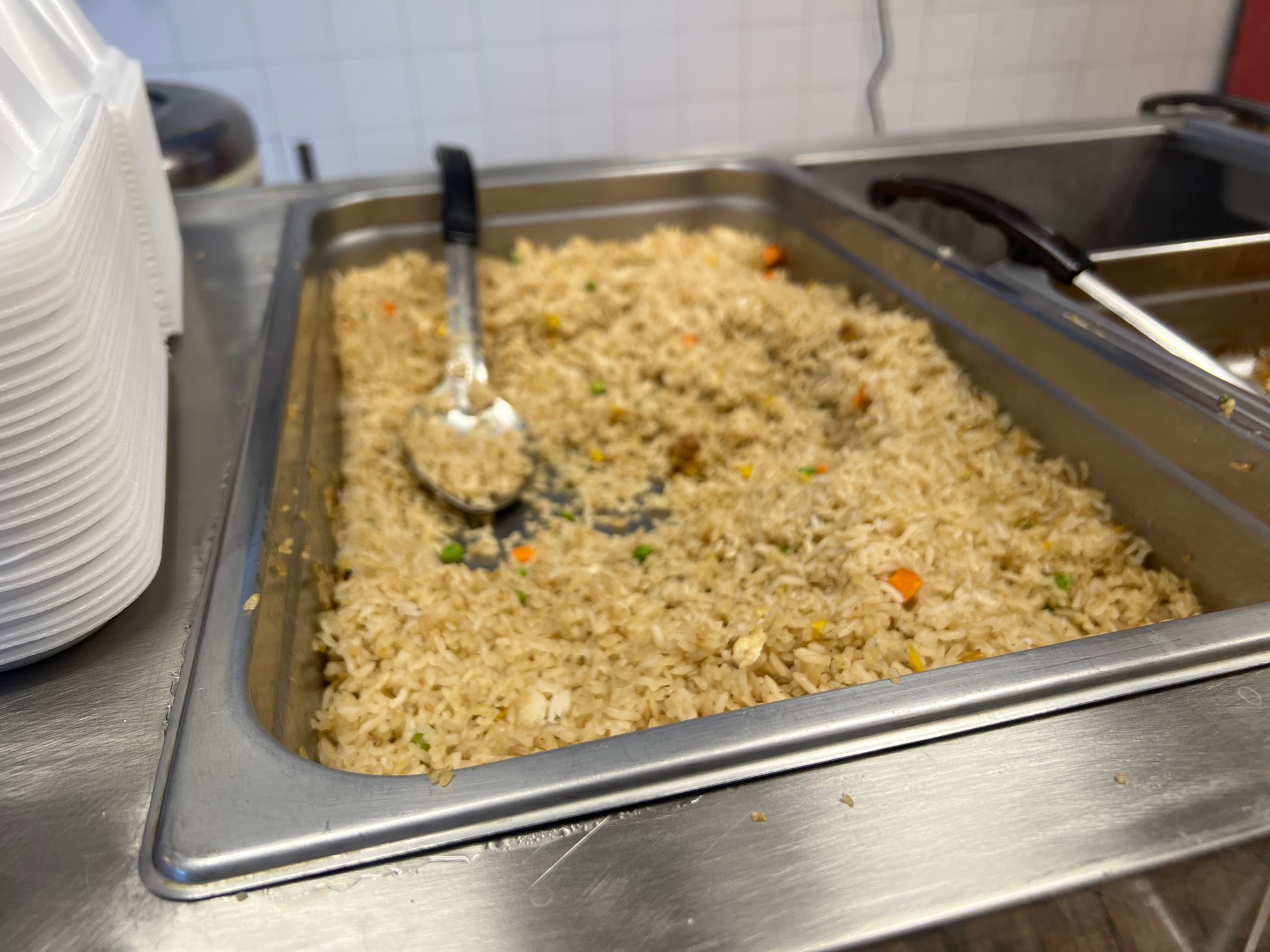 Behind a glass, there is a big metal pan of fried rice. There are small diced carrots, peas, and egg. The serving spoon is resting in the rice with some rice on top. Photo by Alyssa Buckley.