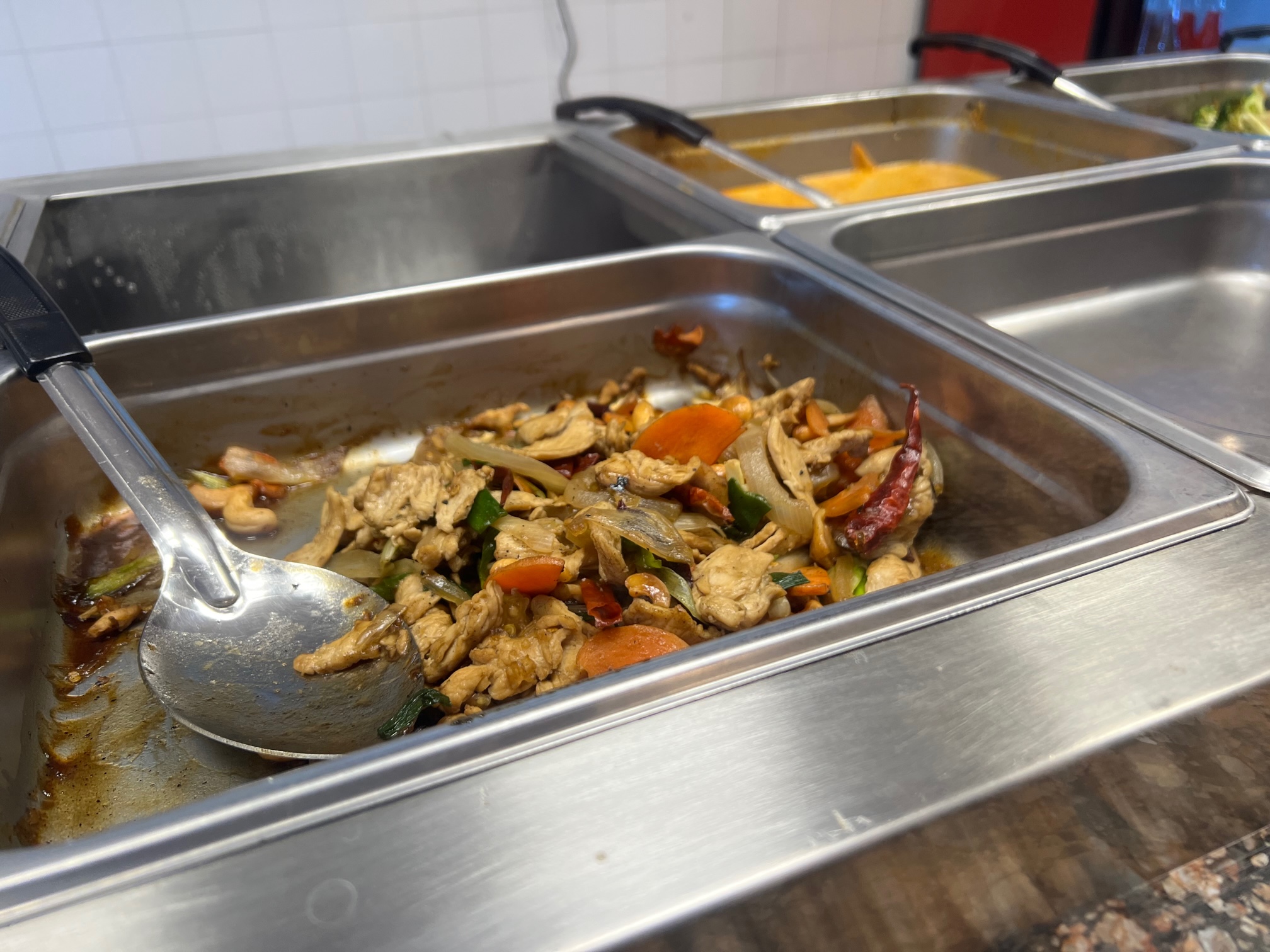 Behind a glass, there is a mostly empty silver pan of chicken with big red chilis and a large serving spoon. Photo by Alyssa Buckley.