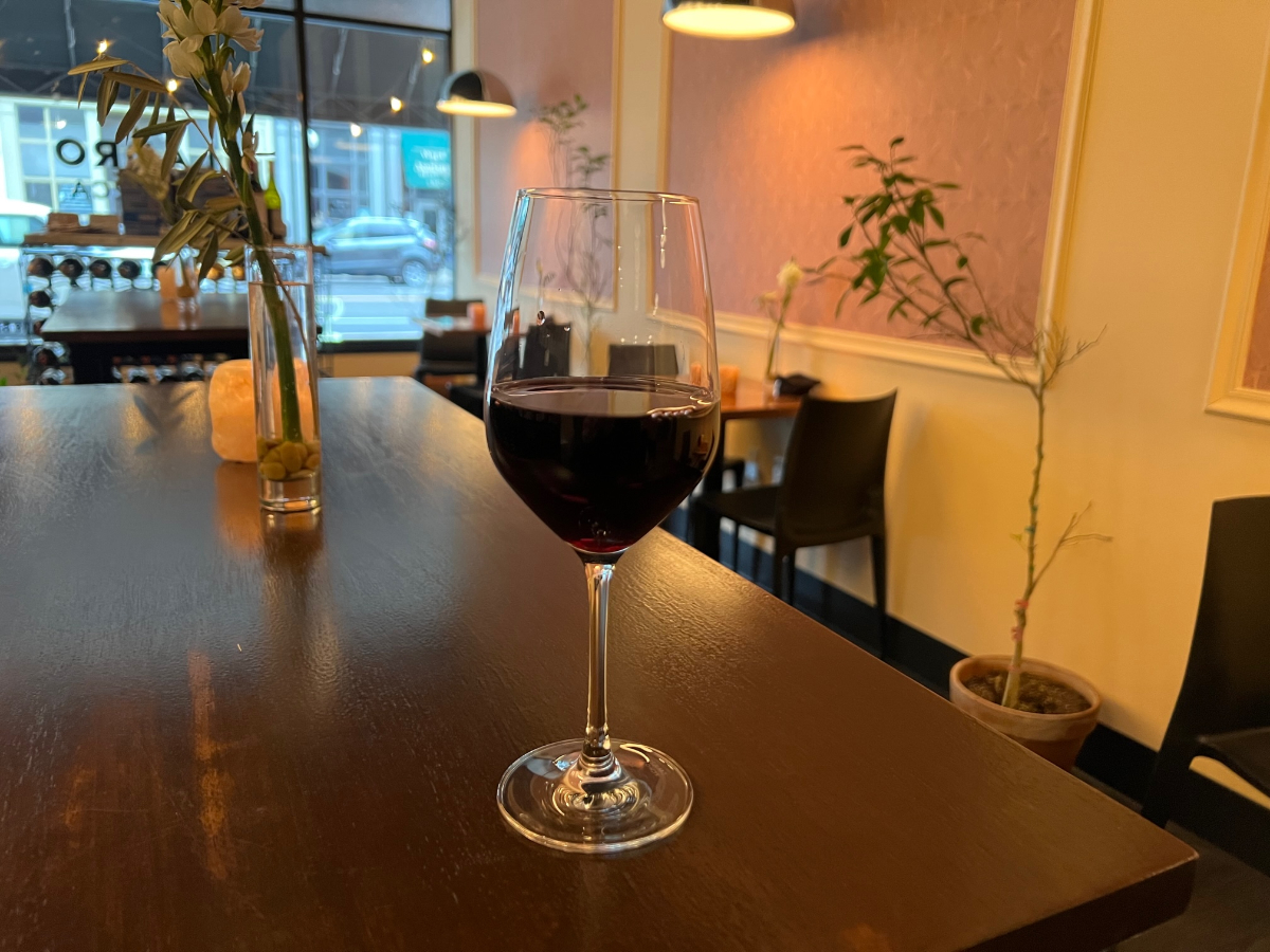 On a brown table, there is a wine glass with red wine inside Ladro Enoteca. There are empty tables for two with simple flowers and branches on the tables. Photo by Alyssa Buckley.