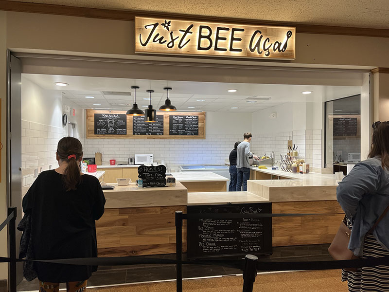 A view of Just Bee AÃ§aÃ­â€™s restaurant from the hallway of the Illini Union. A glowing sign says â€œJust Bee AÃ§aÃ­â€ in a curly, faux-script font. Two employees are at work in the far corner of the clean, brightly-lit kitchen with accents of pine. White subway tile covers the walls and chalk signs list out the menu. Two people wait in line with their backs turned towards the camera. Photo by Julia Freeman.