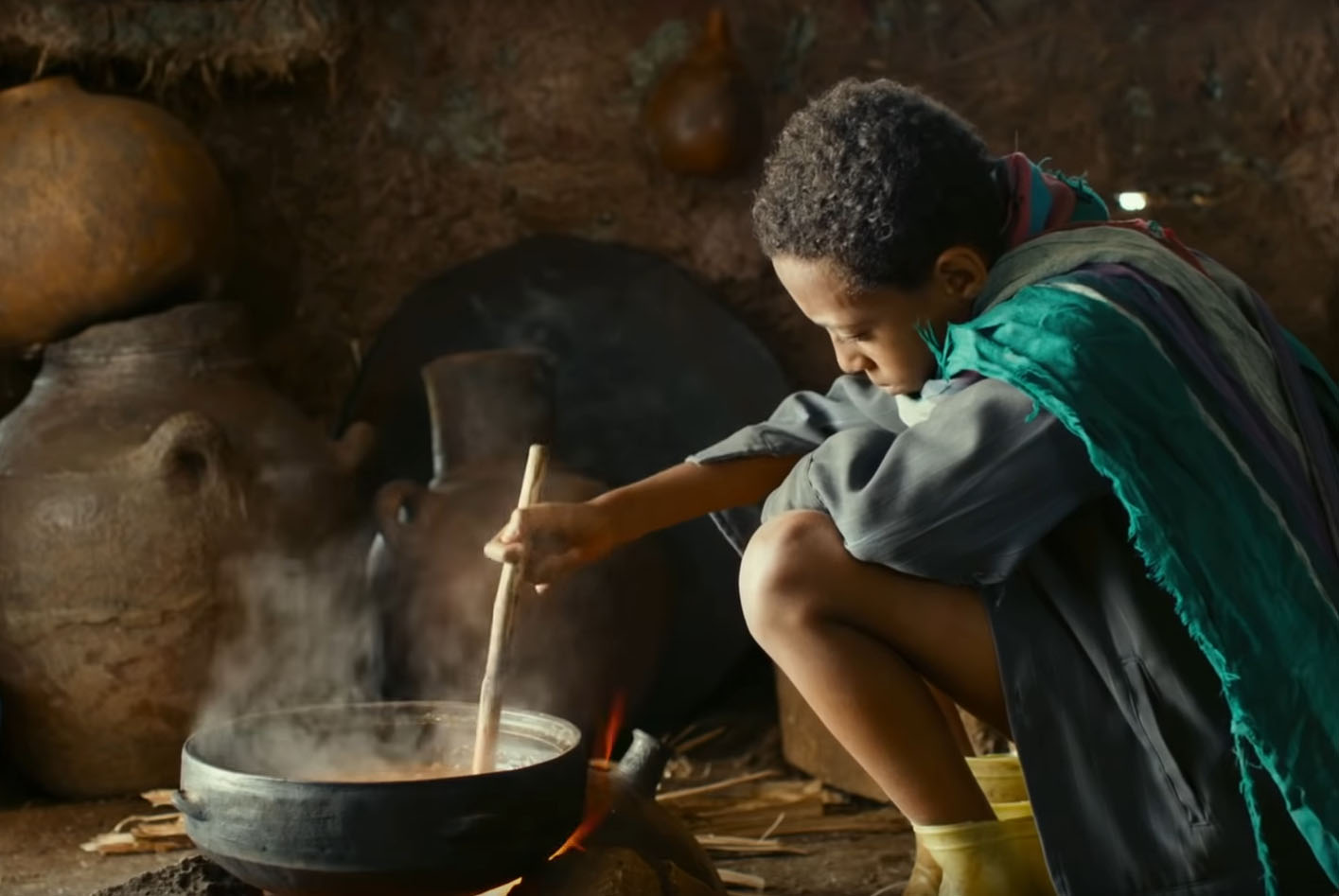 In the movie Lamb, a Black boy sits a black pot with a wooden spoon in a dimly lit room. Photo by Kimstim Films.