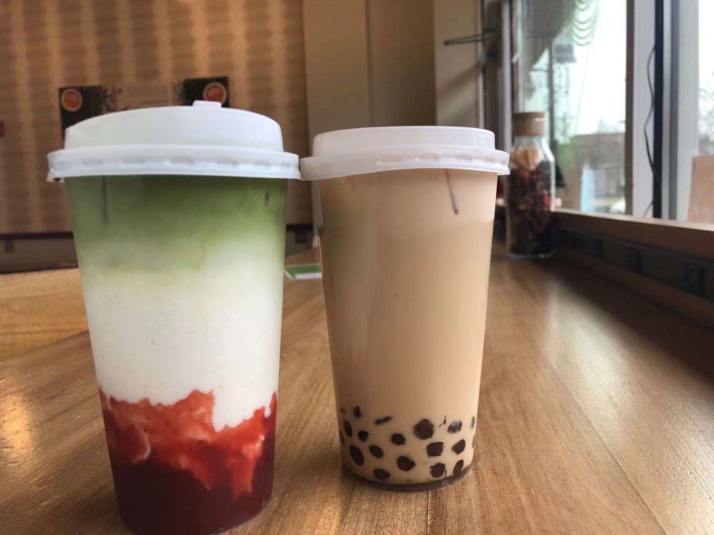 On a wooden table, there are two boba tea drinks in to go plastic cups with white plastic coffee covers. Photo by Xiaohui Zhang.